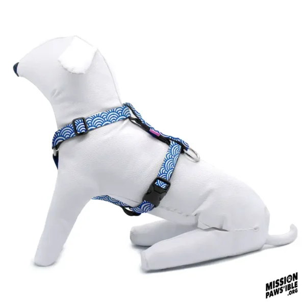 A white dog mannequin wearing the Rollin In The Deep Adjustable Harness, featuring a blue wave pattern, is sitting on a white surface with a "Mission Paws'ible" logo in the corner.