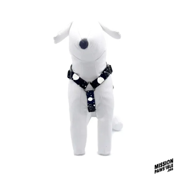 A plush white dog is sitting upright on a white background, wearing the Into The Cosmos Adjustable Harness, which features a black design with white spots. The Mission Paws'ible logo is positioned in the bottom right corner.