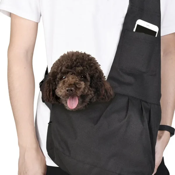 A small brown dog with curly fur peeks out from a 'Mums The Word' Puppy Pack, a black sling carrier worn by a person. The person's phone sits in the pack's pocket.