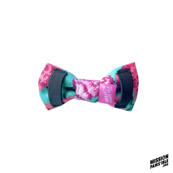 A vibrant pink and green bow tie named "Flower Power Aqua Bow Tie" flaunts the text "Mission Pawsible" and comes with two black elastic bands on the sides.