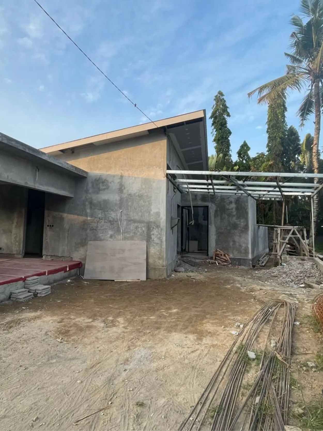 Partially constructed building with concrete walls, metal rods, and scattered construction materials. Palm trees and a clear sky are in the background.