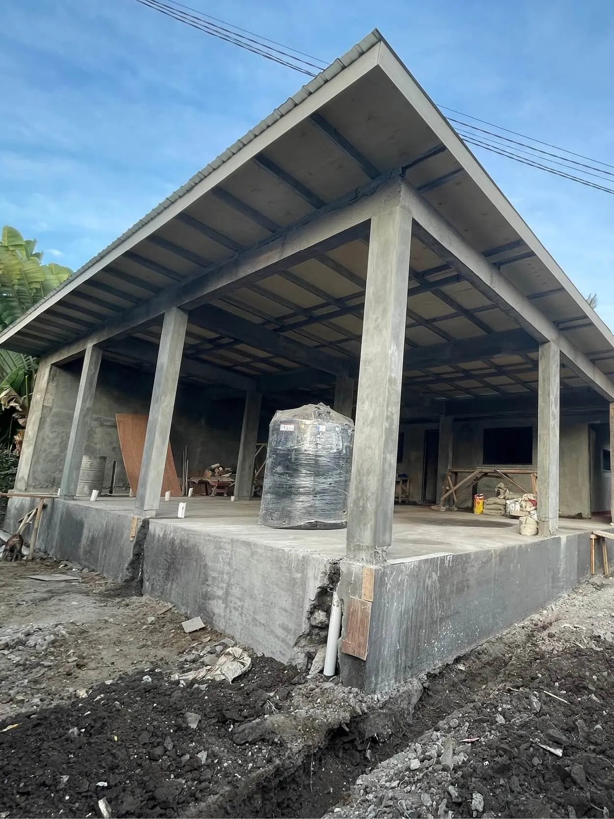 A partially constructed building with a corrugated metal roof, concrete pillars, and an unfinished interior. The ground is uneven and a large black barrel is on the concrete floor.