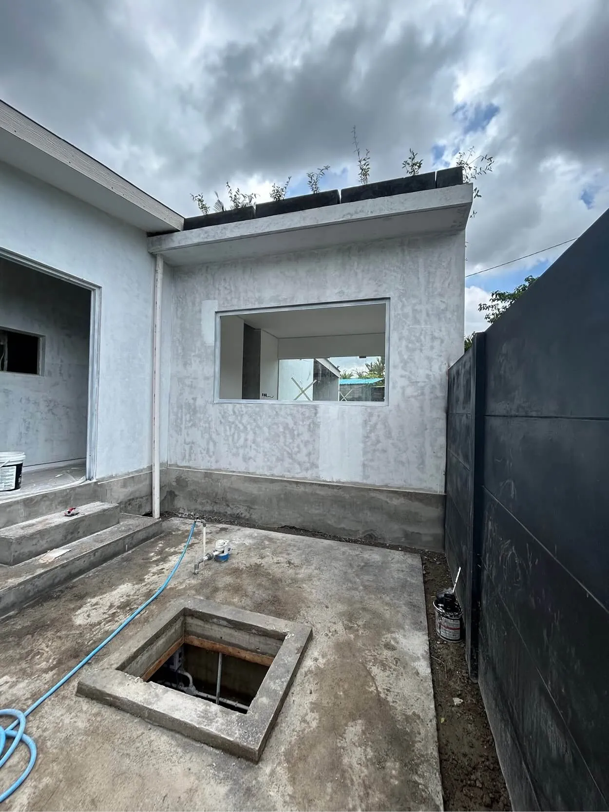 A partially constructed building with an unfinished wall and an open square pit in the concrete ground. Steps lead up to a doorway on the left. Overcast sky in the background.