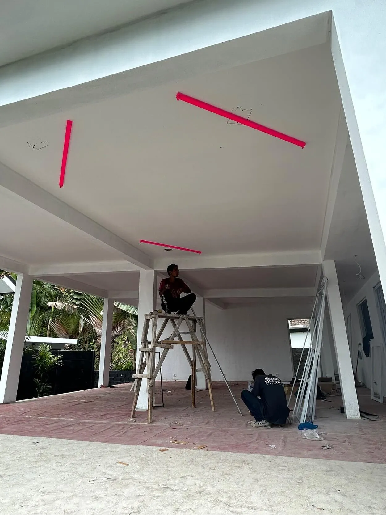 Two people work on an unfinished porch. One sits on scaffolding under a white ceiling with red strips. Another is on the ground near tools and materials.