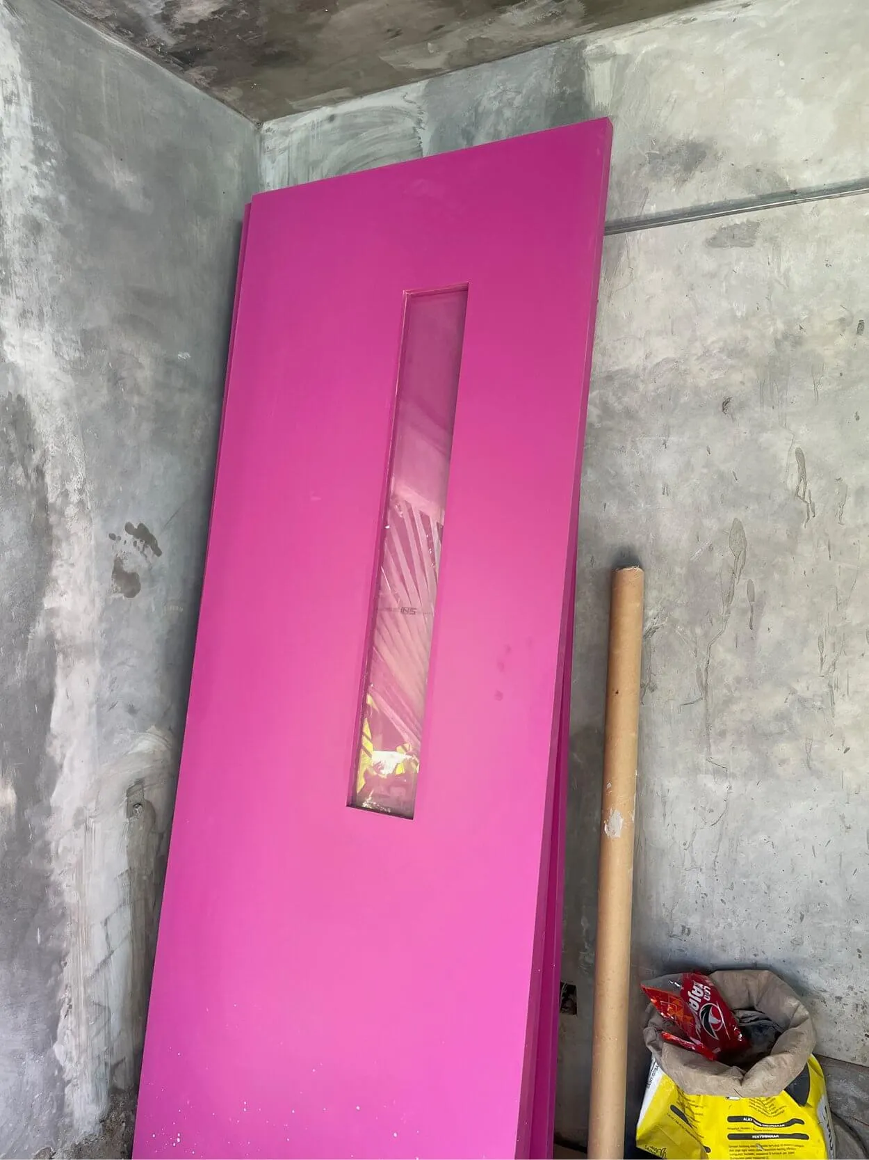 A vibrant pink door with a narrow vertical window leans against a gray cement wall in a corner. Nearby are a wooden rod and a yellow bag on the floor.