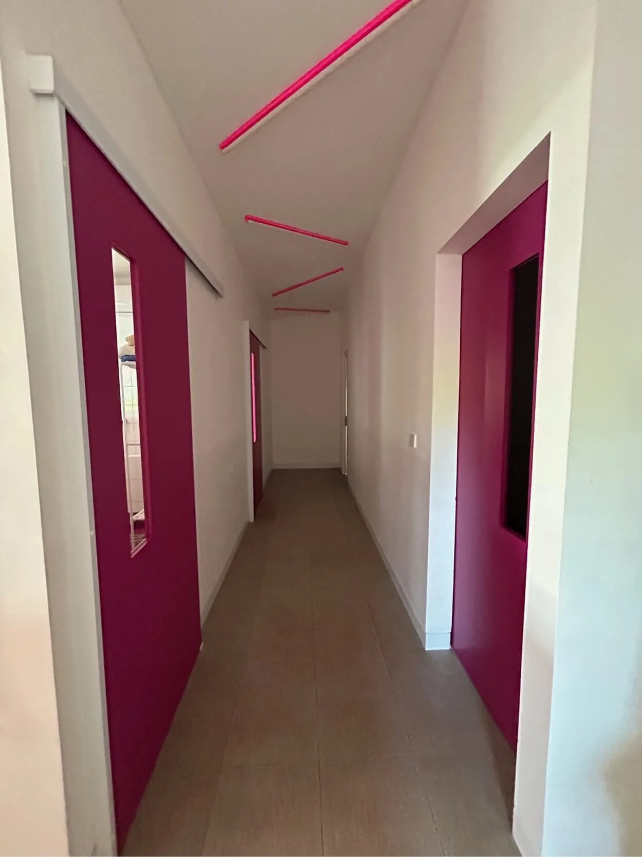 A hallway with pink sliding doors and red ceiling lights, leading to an open doorway. Walls and floor are white and beige.