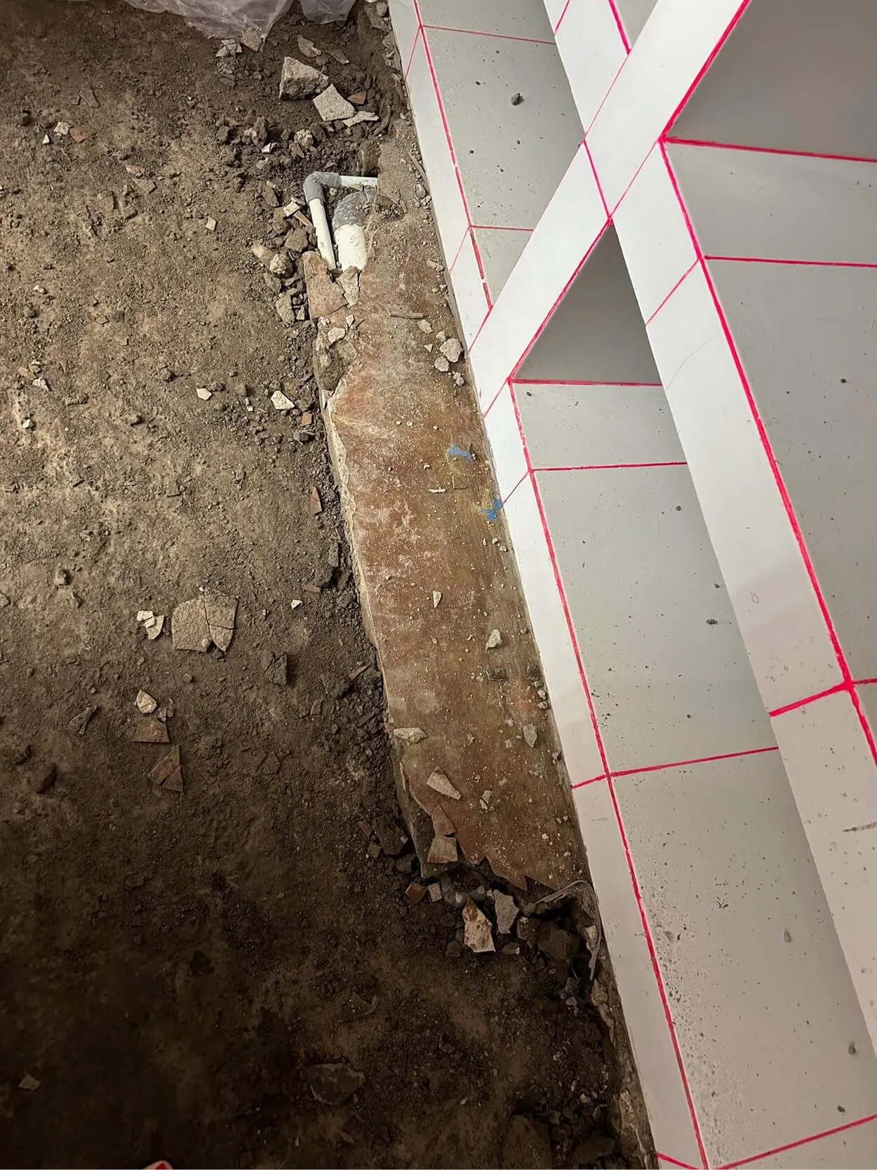 Construction site showing a dirt floor, broken concrete slab, pipes, and a wall with tiled sections marked by pink guidelines.