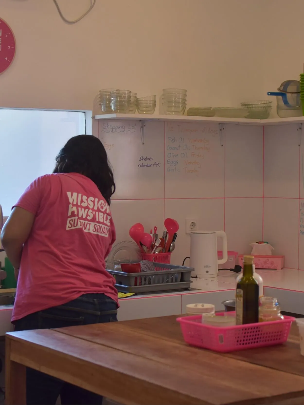 Person in a pink shirt standing in a kitchen, facing the counter with dishes and a kettle. Various kitchen items are placed on the countertop and shelves.