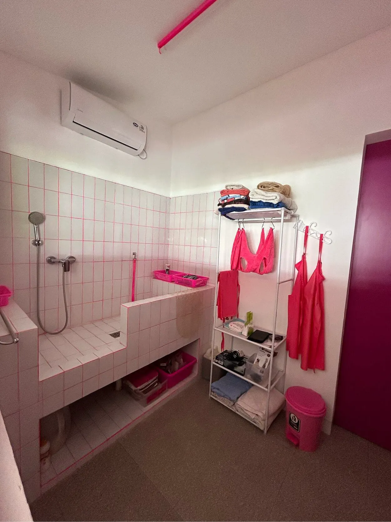 Bathroom with a tiled shower area, a metal shelf with pink towels and clothes, a pink trash bin, and an air conditioning unit on the wall.