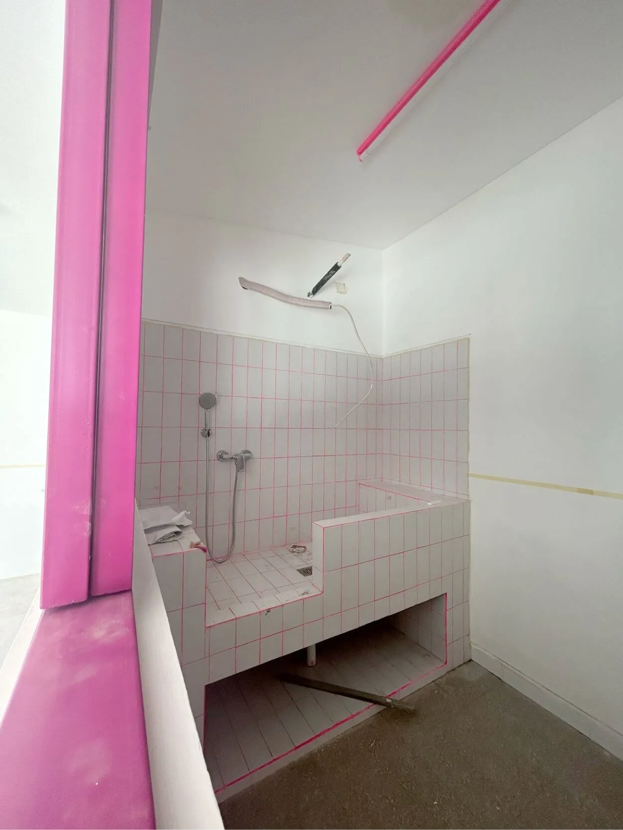 Bathroom with white tiles and pink grout, featuring a shower area and a broken step. Exposed pipes and a fluorescent light are visible.