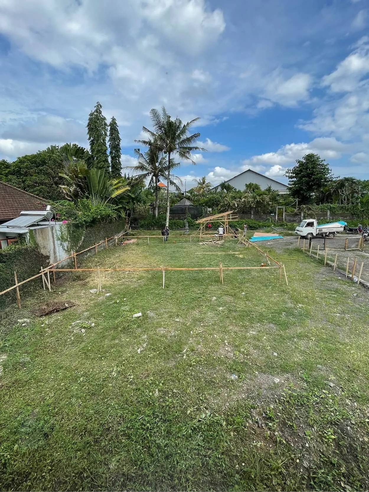 Grassy field with wooden markers outlining a rectangle, surrounded by trees and small buildings under a partly cloudy sky.