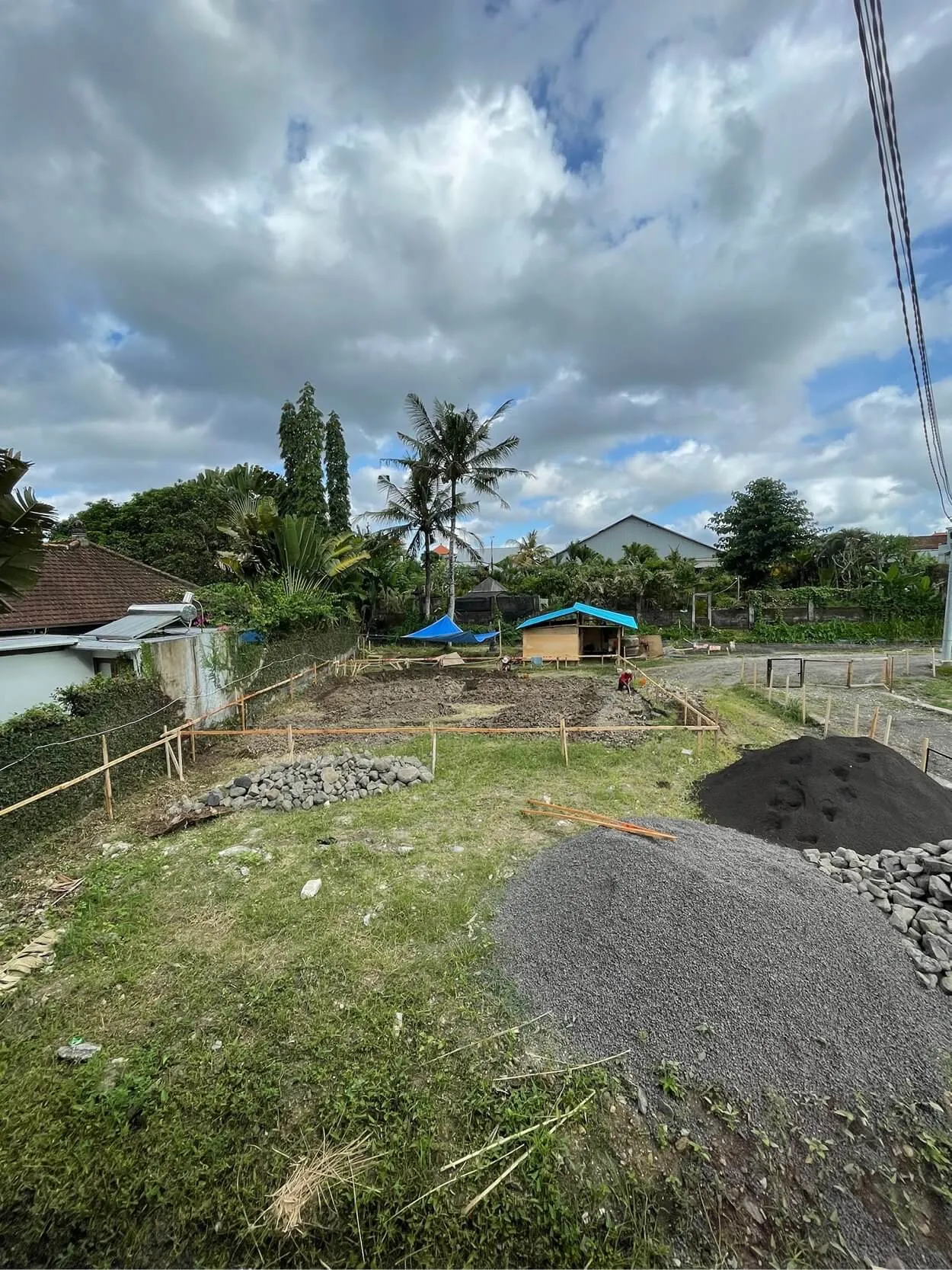 A grassy construction site with gravel piles and wooden stakes. Two blue tents are set up near the center. A palm tree and several houses are visible in the background under a cloudy sky.