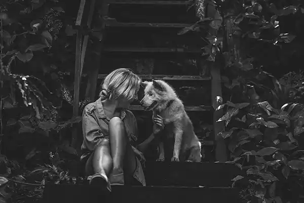 A person sits on outdoor steps, affectionately interacting with a dog, surrounded by dense foliage.